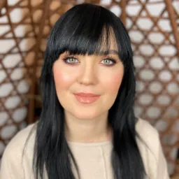 Portret kobiety o zielonych oczach z czarnymi włosami i delikatnym, naturalnym makijażu w odcieniach brzoskwini i beżu, na tle ażurowego, wiklinowego fotela.