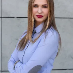 Portret kobiety w błękitnej koszuli z szarymi łatami na łokciach, skrzyżowane ramiona, ciemna szminka, na tle szarej ściany z poziomymi liniami.