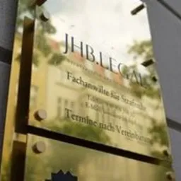 Złota tabliczka informacyjna z napisem 'JHB.LEGAL Fachanwälte für Strafrecht' przy wejściu do biura w Berlinie, widoczna z boku, z refleksami światła.