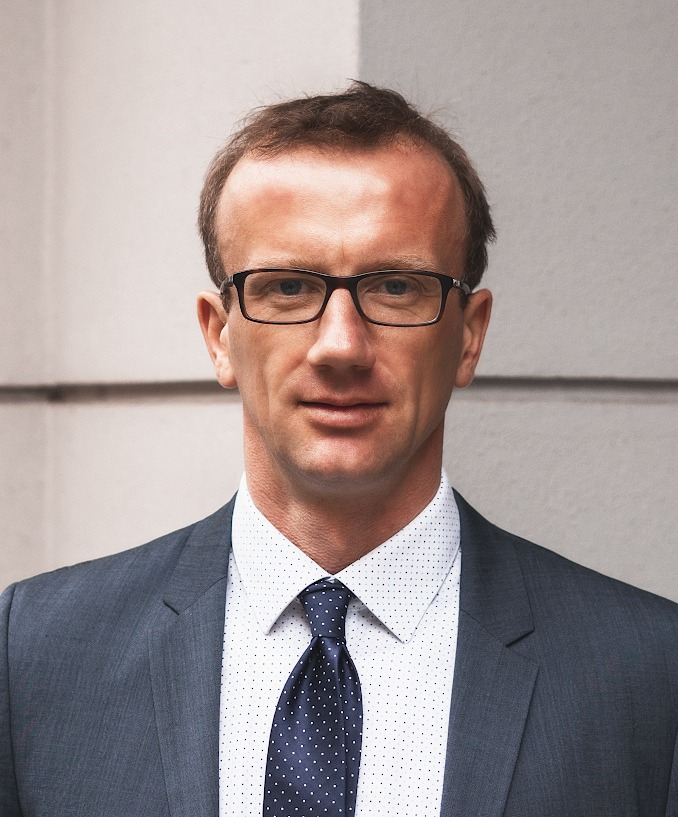 Portret mężczyzny w garniturze, białej koszuli w drobne kropki i granatowym krawacie w białe kropki, w okularach, na tle jasnej ściany.