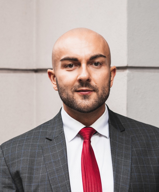 Portret mężczyzny w garniturze w kratę i czerwonym krawacie na tle jasnej ściany.