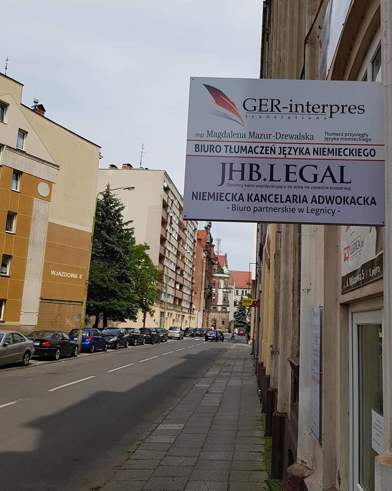 Tablica informacyjna na budynku z napisem 'GER-interpres translations, Biuro Tłumaczeń Języka Niemieckiego' oraz 'JHB.LEGAL Niemiecka Kancelaria Adwokacka' na tle ulicy w Legnicy.