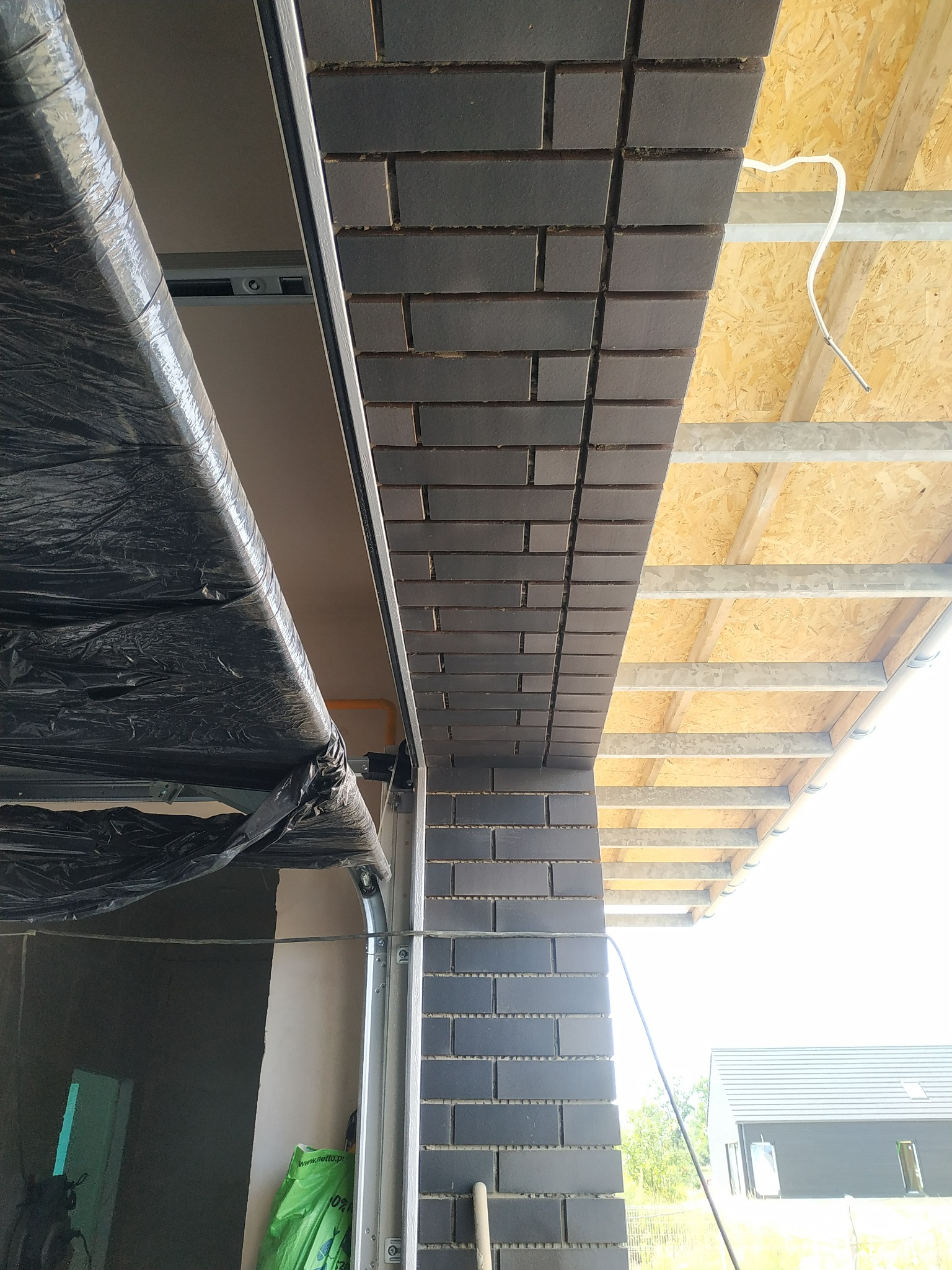 Fragment elewacji z ciemnej cegły klinkierowej w trakcie montażu, widoczne rusztowanie i konstrukcja dachu, boczny widok na bramę garażową z folią ochronną.