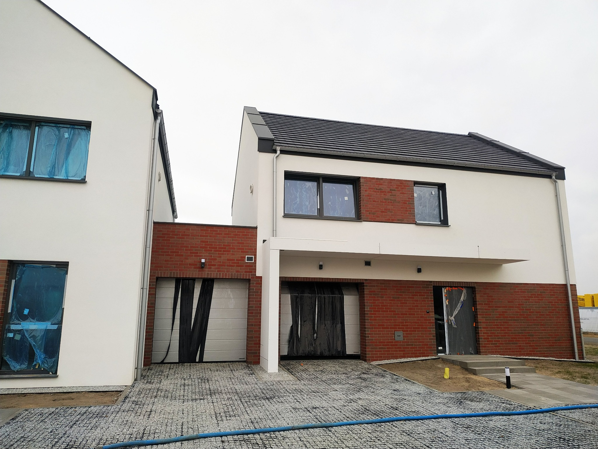 Dwa nowoczesne domy w trakcie budowy, z białymi elewacjami i fragmentami wykończonymi czerwoną cegłą, okna zabezpieczone folią, a garaże osłonięte czarną folią malarską, kostka brukowa...