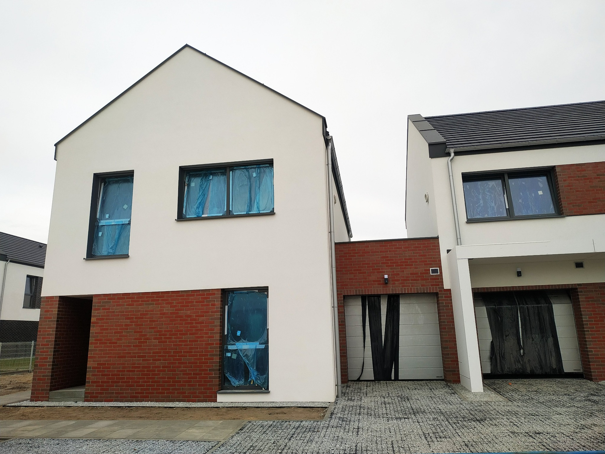 Nowoczesny dom w trakcie budowy z białymi ścianami, czerwoną cegłą na elewacji dolnej części i oknami zabezpieczonymi folią ochronną. Podjazd wyłożony kostką.