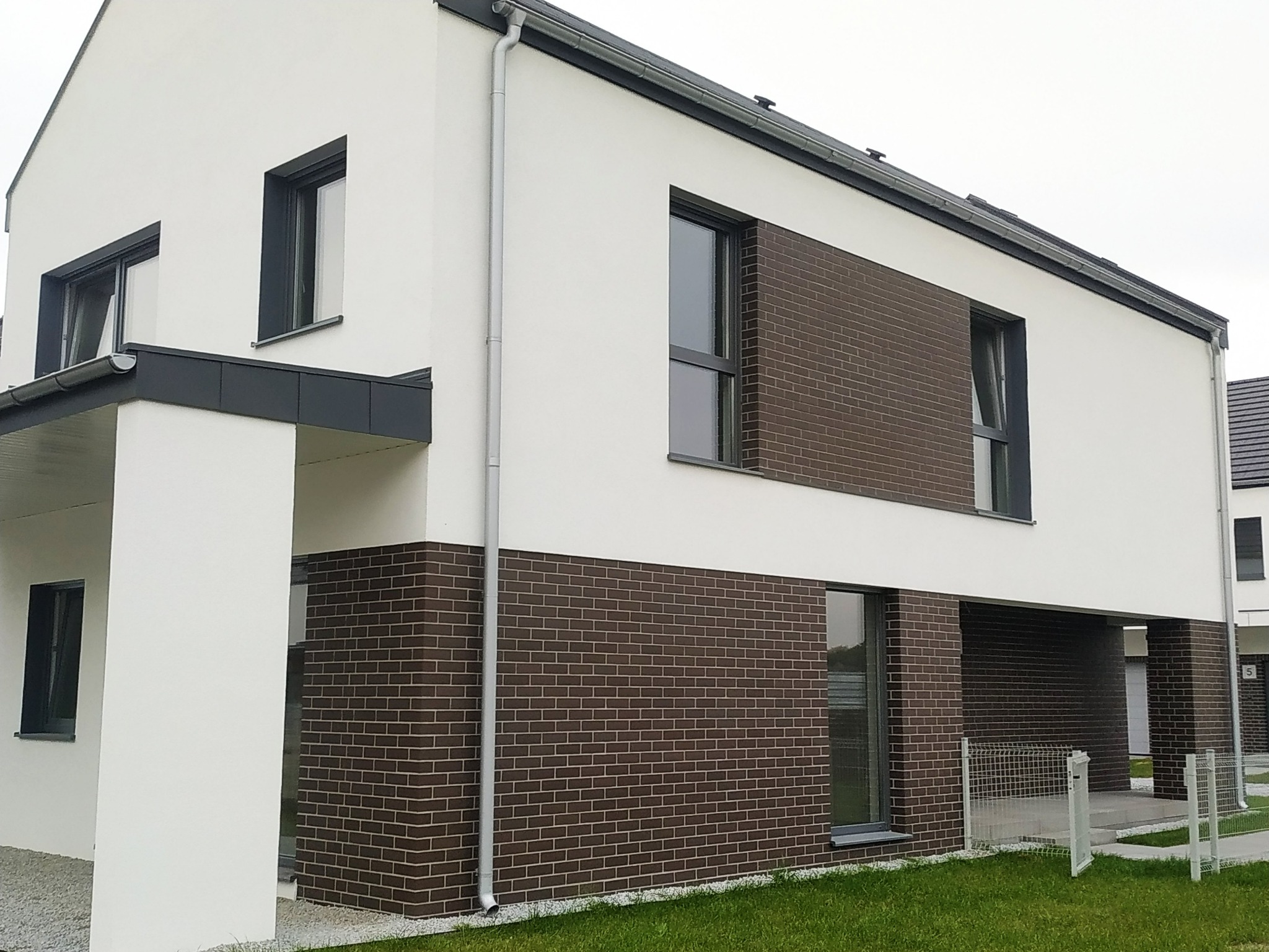 Nowoczesny dom jednorodzinny z białą elewacją i fragmentami wykończonymi brązową cegłą klinkierową, z widocznymi oknami w ciemnych ramach i szarą rynną.