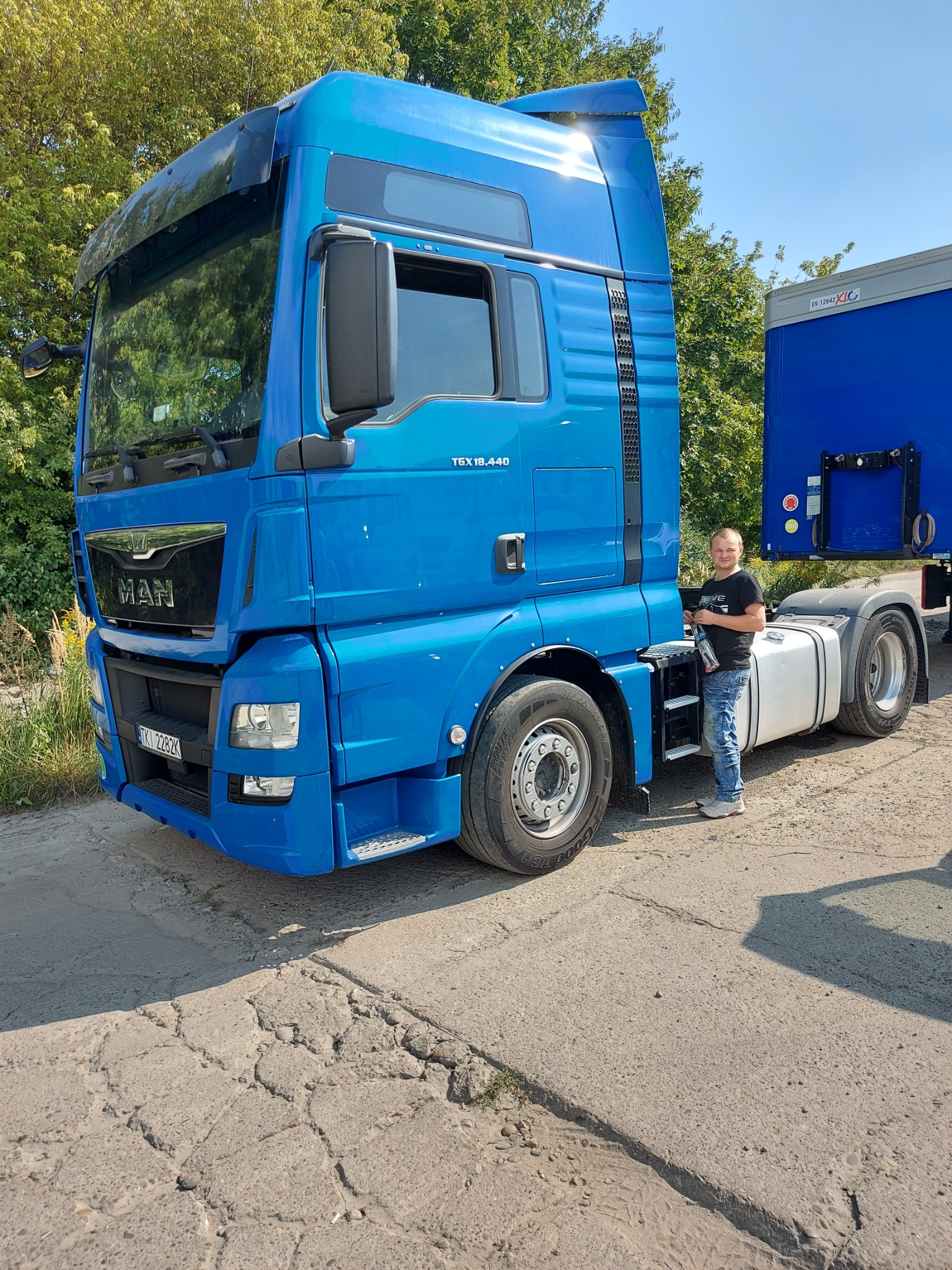 Niebieski ciągnik siodłowy MAN TGX 18.440 z kierowcą stojącym przy kabinie, na tle zieleni i częściowo widocznej naczepy.