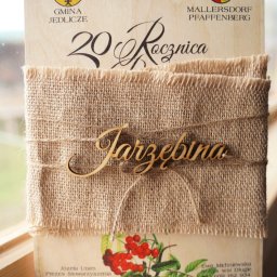 Form-Art Pracowania Atystyczna - Drewniane zaproszenie na 20. rocznicę z herbami Gminy Jedlicze i Mallersdorf Pfaffenberg, przewiązane jutowym materiałem i sznurkiem z drewnianym napisem 'Jarzębina', ozdobione motywem jarzębiny.