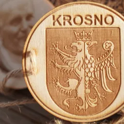 Drewniany, okrągły brelok z wygrawerowanym herbem Krosna na tle drewnianego pudełka i fragmentu okrągłej drewnianej ozdoby z wizerunkiem osoby.