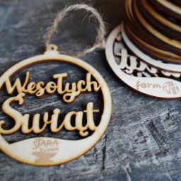 Form-Art Pracowania Atystyczna - Drewniane ozdoby świąteczne z napisem 'Wesołych Świąt' i logo firmy na szarym, teksturowanym tle, jeden wisi na sznurku, a reszta ułożona w stos.