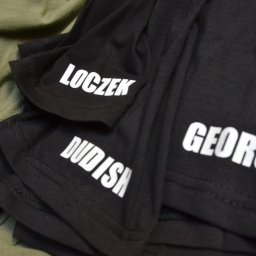 Form-Art Pracowania Atystyczna - Stos czarnych koszulek z białymi napisami 'LOCZEK', 'DUDISH' i 'GEORGE' na rękawach oraz na przodzie, leżących na zielonej tkaninie.
