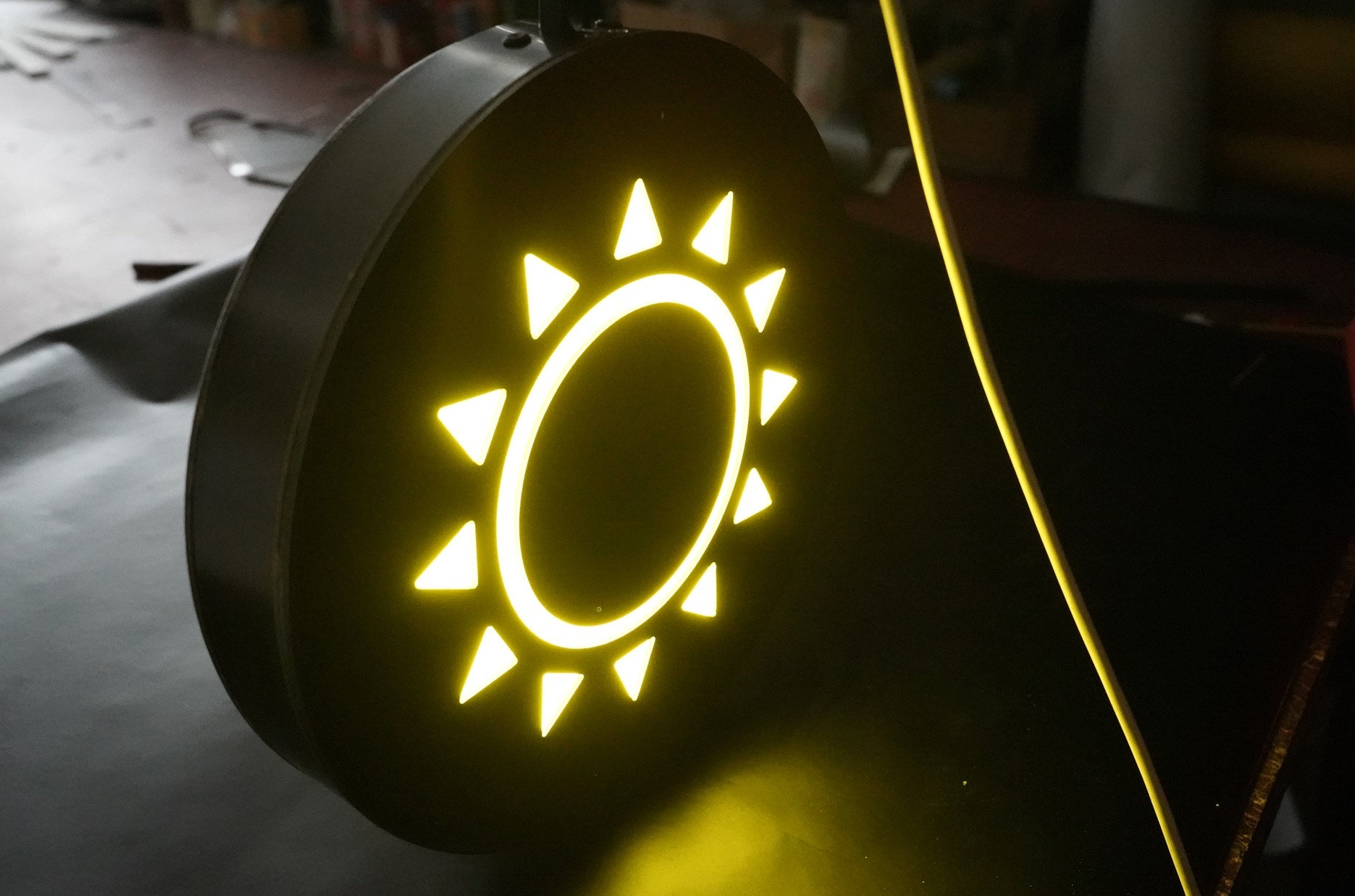 Podświetlany, okrągły szyld z motywem słońca wykonany z żółtego neonu, umieszczony na ciemnym tle.