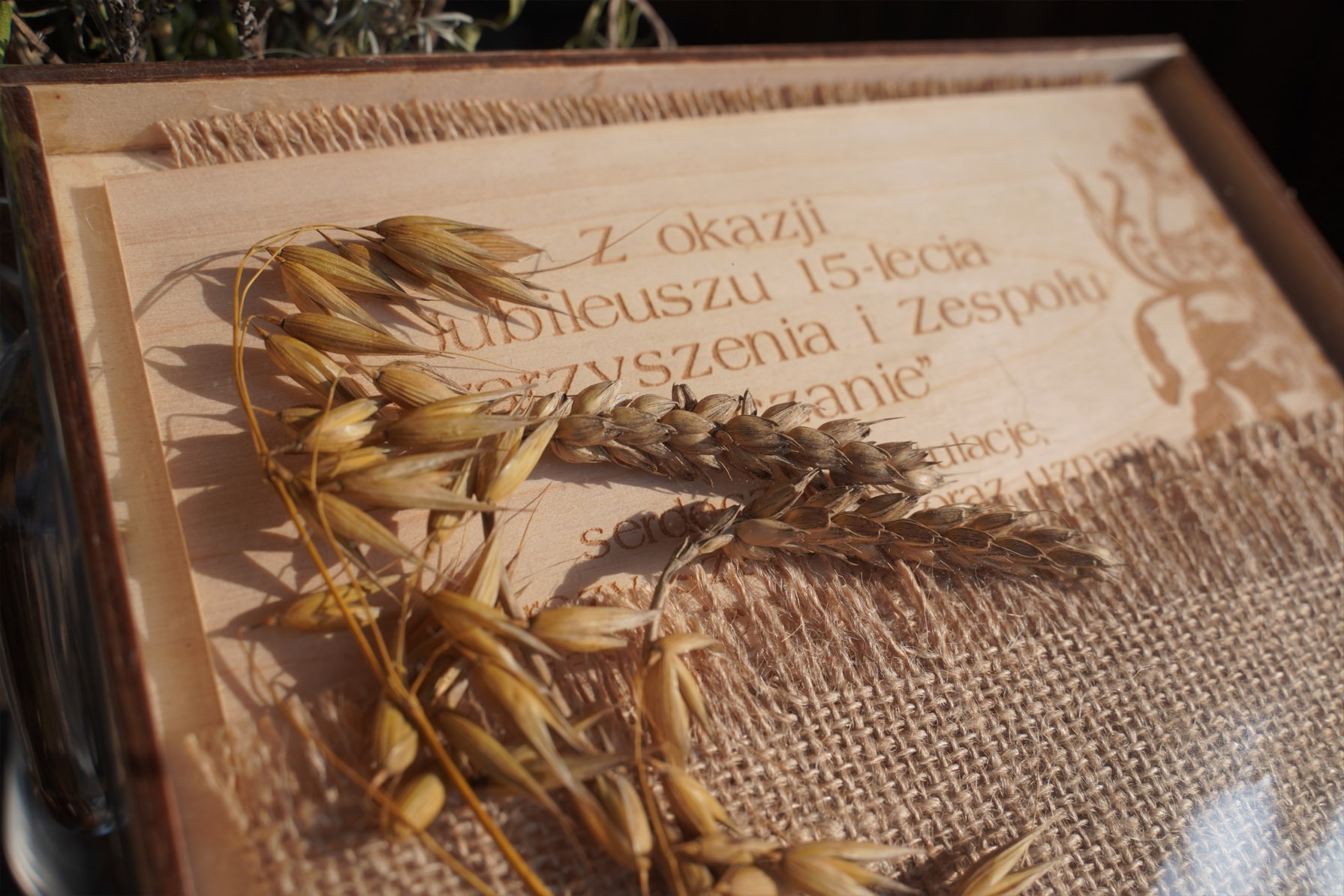 Grawerowana drewniana tablica z tekstem 'Z okazji Jubileuszu 15-lecia...' i motywem roślinnym, zdobiona jutowym płótnem i kłosami zbóż.