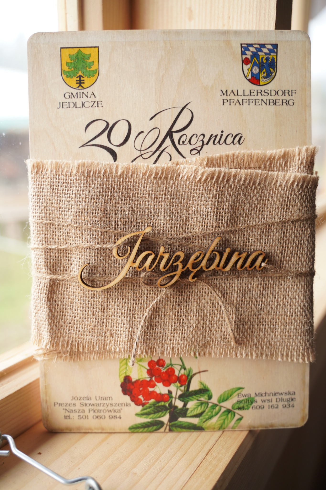 Drewniane zaproszenie na 20. rocznicę z herbami Gminy Jedlicze i Mallersdorf Pfaffenberg, przewiązane jutowym materiałem i sznurkiem z drewnianym napisem 'Jarzębina', ozdobione motywem jarzębiny.