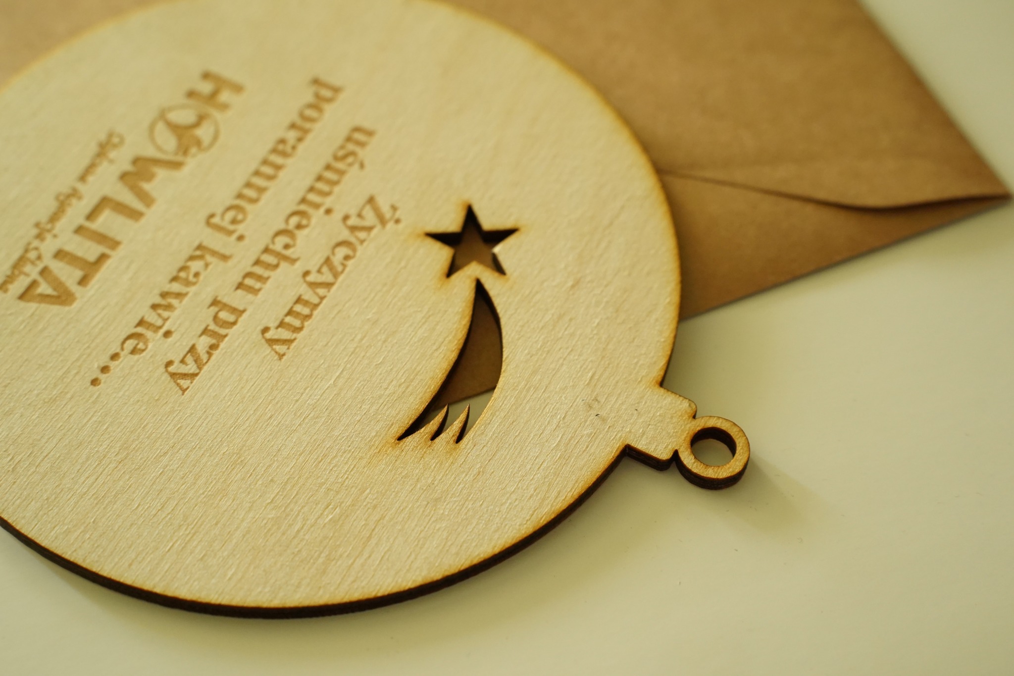 Drewniana zawieszka w kształcie bombki z wygrawerowanym tekstem 'Życzymy uśmiechu przy porannej kawie...' i logiem firmy 'HOWLITA', na tle koperty.