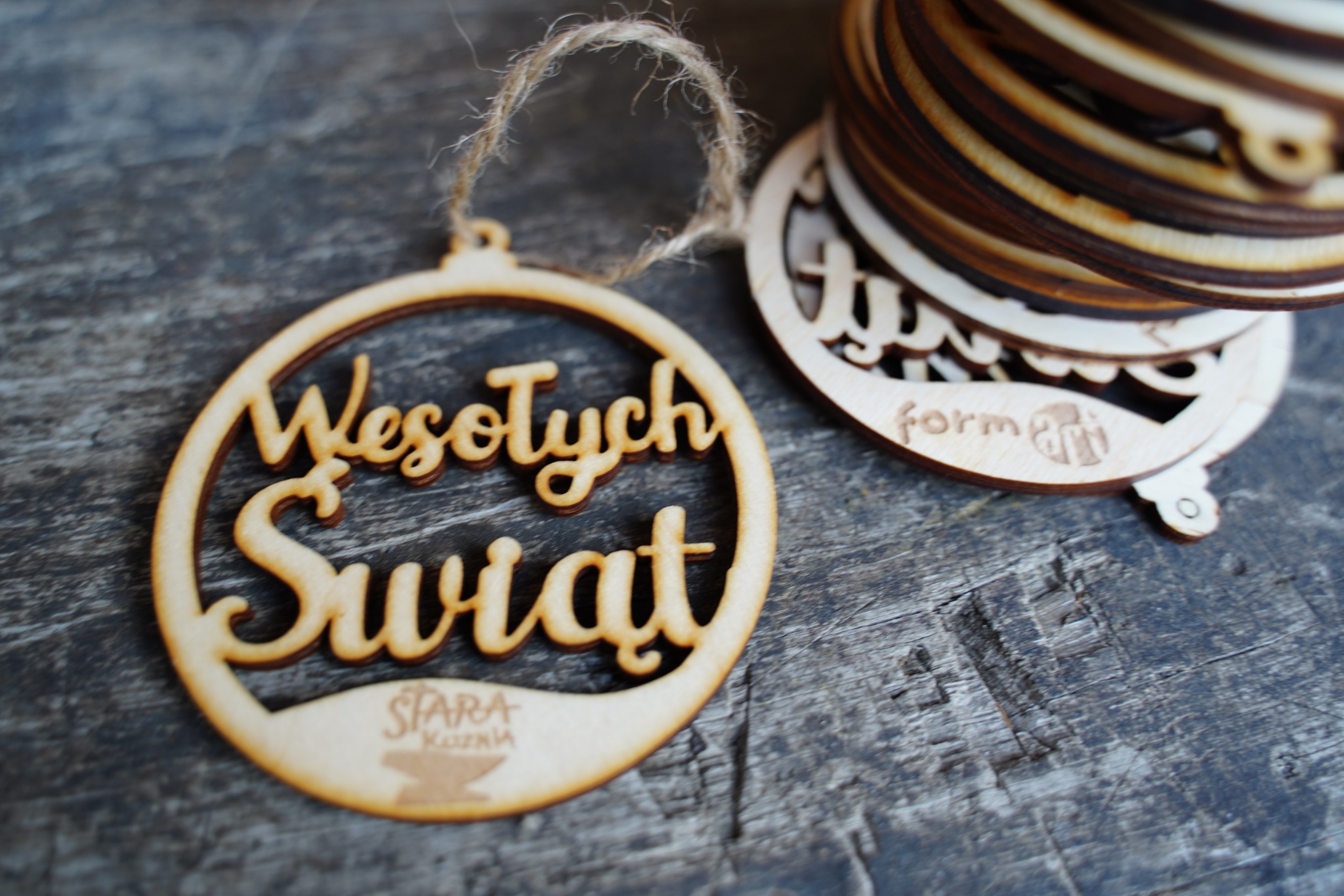 Drewniane ozdoby świąteczne z napisem 'Wesołych Świąt' i logo firmy na szarym, teksturowanym tle, jeden wisi na sznurku, a reszta ułożona w stos.