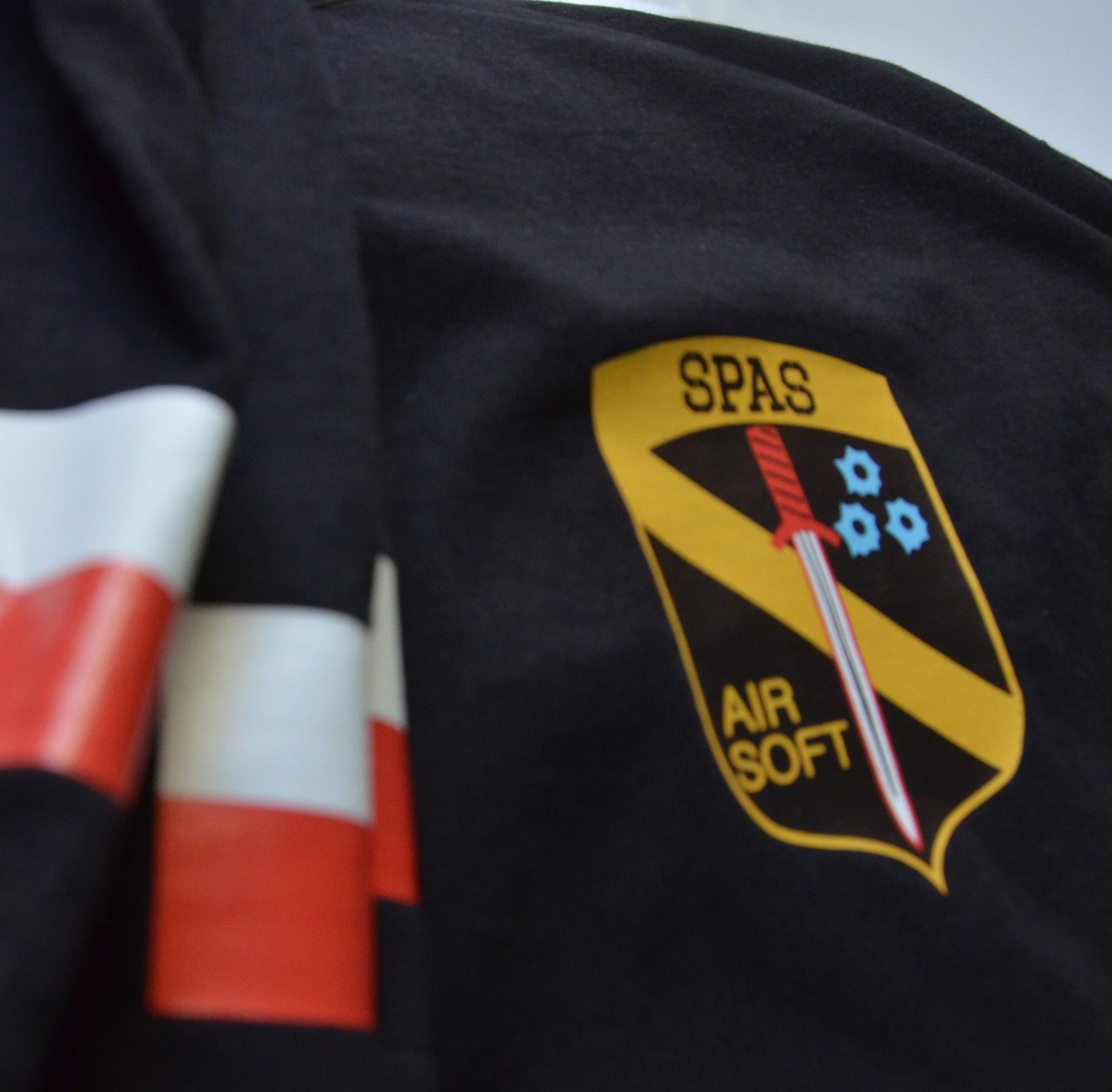 Czarne ubranie z naszywką SPAS Air Soft z motywem miecza i śladów po kulach, z fragmentem biało-czerwonej flagi w tle.