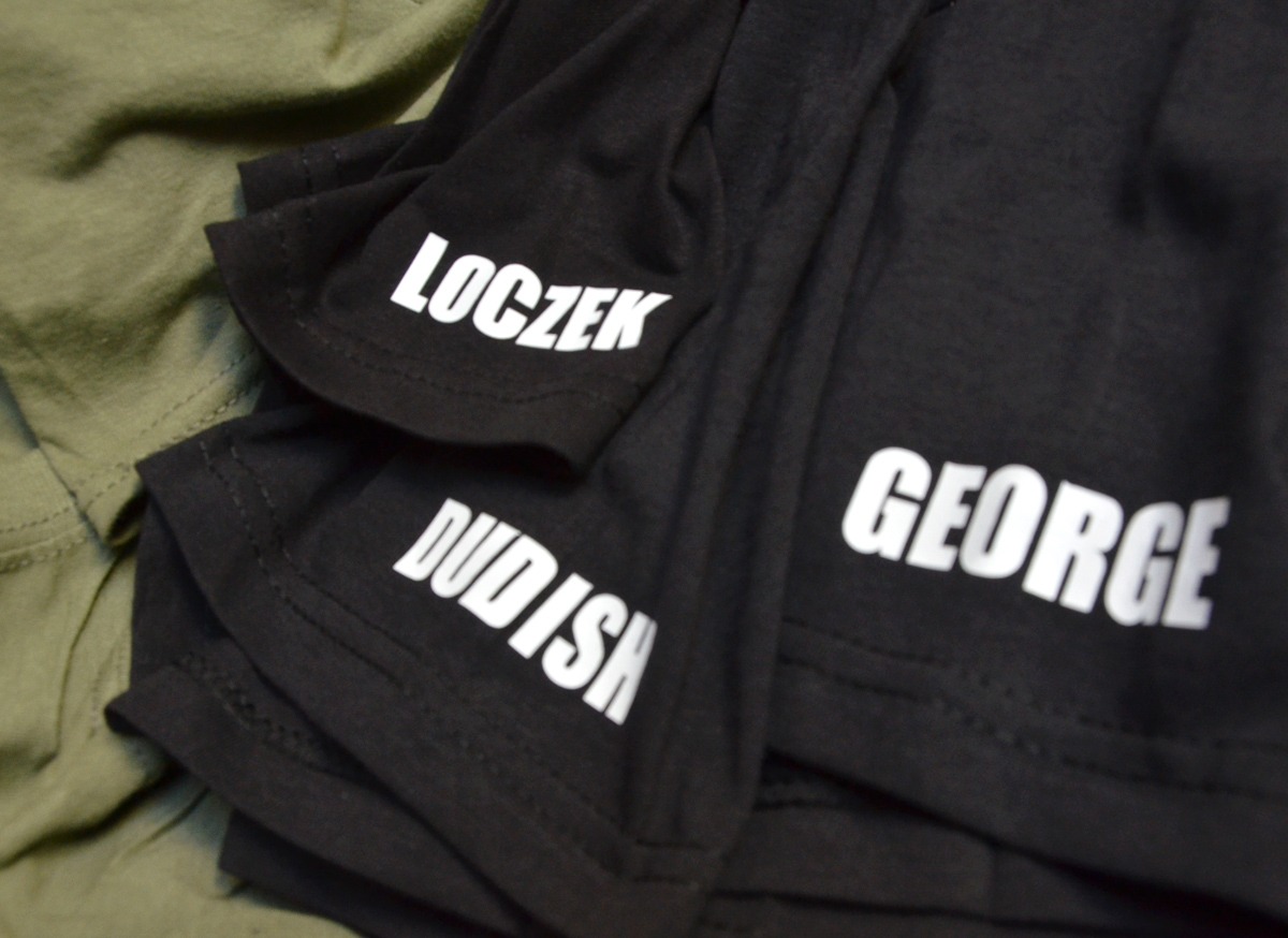 Stos czarnych koszulek z białymi napisami 'LOCZEK', 'DUDISH' i 'GEORGE' na rękawach oraz na przodzie, leżących na zielonej tkaninie.