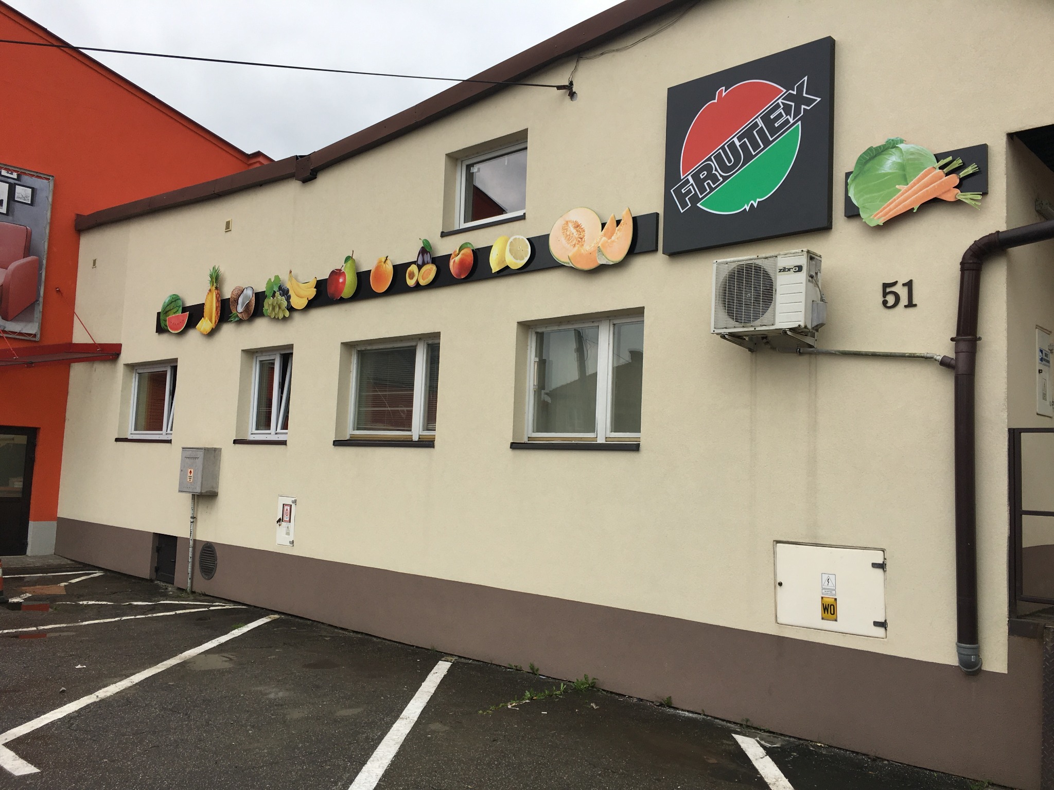 Elewacja budynku z logo Frutex i dekoracyjnymi elementami przedstawiającymi owoce i warzywa, umieszczonymi na czarnym pasku. Obok logo znajduje się klimatyzator i numer budynku '51'.