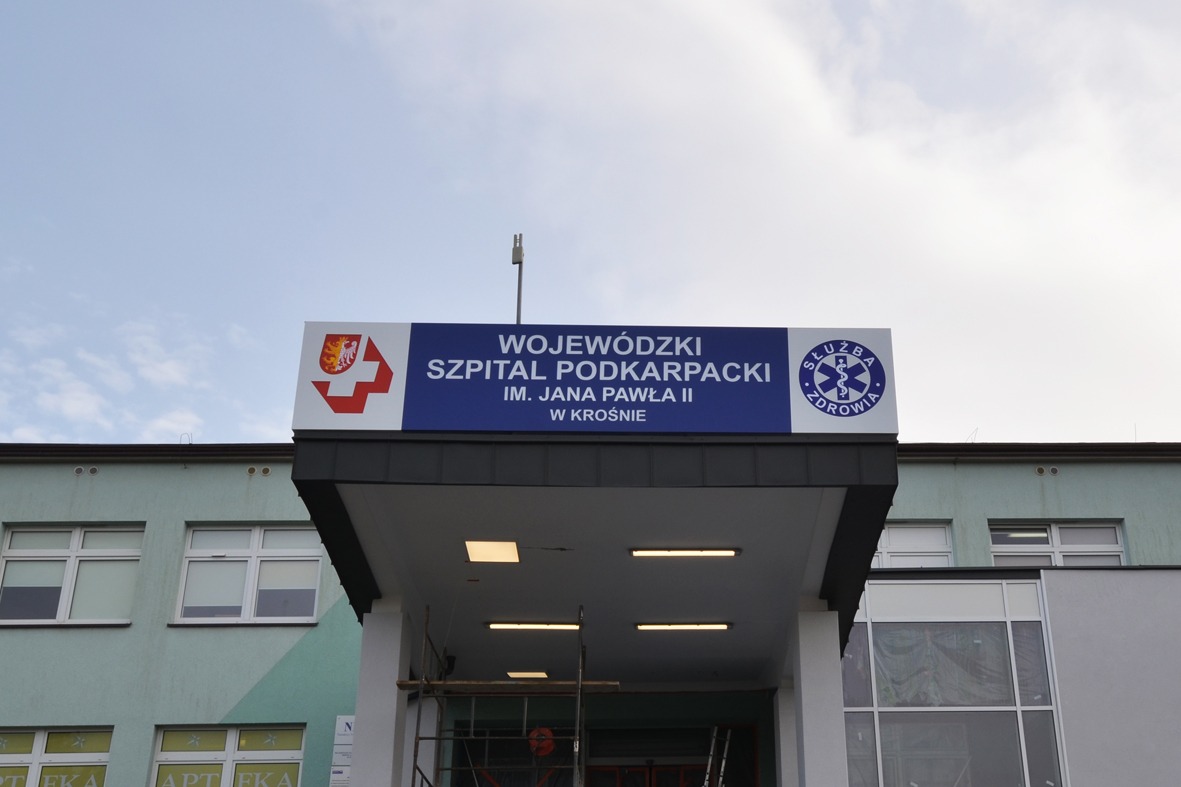 Tablica informacyjna z nazwą Wojewódzkiego Szpitala Podkarpackiego im. Jana Pawła II w Krośnie, umieszczona nad wejściem do budynku. Widoczne logo szpitala i symbol służby zdrowia.
