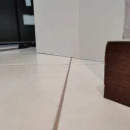 Beżowe płytki podłogowe z brązowymi fugami, białe ściany i ciemna listwa przypodłogowa widziane z niskiej perspektywy.