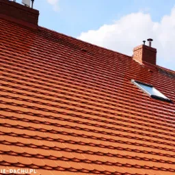 Czysty, mokry dach pokryty czerwoną dachówką karpiówką po umyciu, z widocznymi dwoma oknami dachowymi i ceglanym kominem na tle błękitnego nieba.