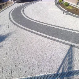 Zakrzywiony podjazd z jasnoszarej kostki z ciemnoszarymi pasami, widoczna studzienka odpływowa i cień osoby w prawym dolnym rogu.