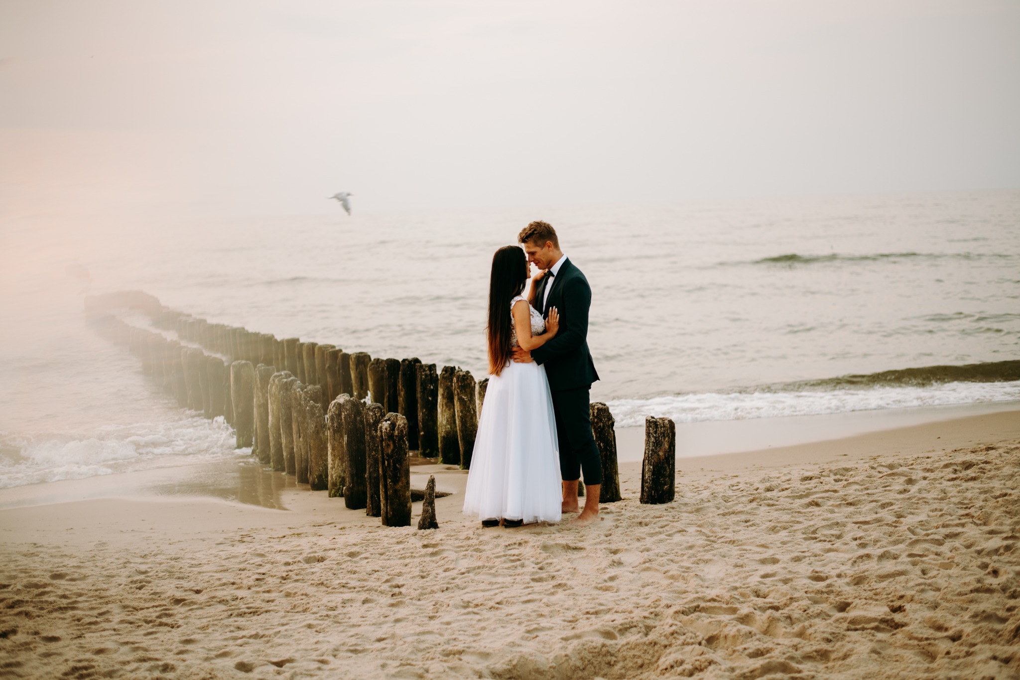 Para młoda obejmująca się na piaszczystej plaży z drewnianymi palami w tle, mewa w locie, stonowana kolorystyka.