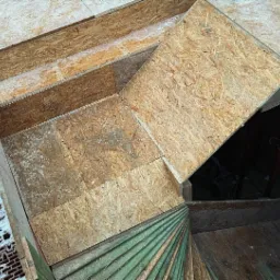 Szkielet poddasza z płyt OSB w trakcie ocieplania, widoczne fragmenty muru z pustaków ceramicznych i zielonej wełny mineralnej, oświetlenie robocze w tle.