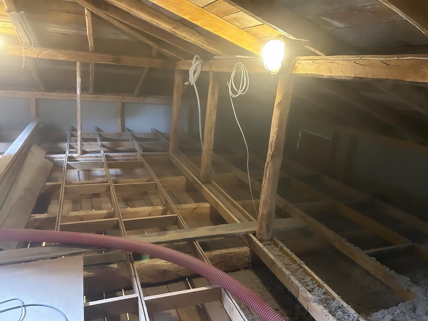 Konstrukcja drewniana poddasza z częściowo wypełnioną izolacją, widoczna lampa budowlana i czerwony wąż do wdmuchiwania materiału izolacyjnego.