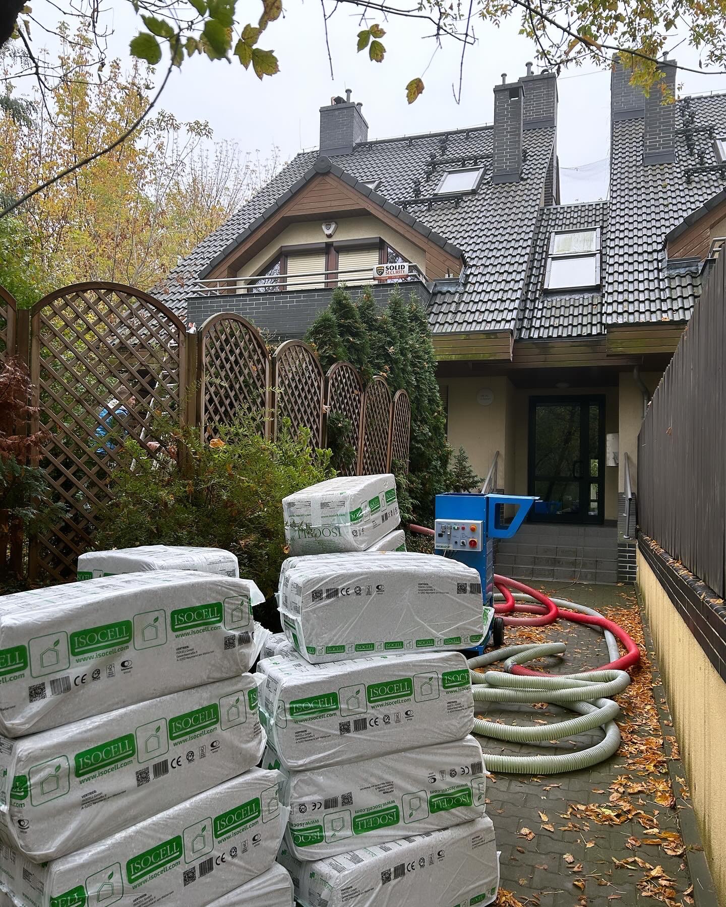 Stos worków z wełną izolacyjną Isocell i maszyna do wdmuchiwania, przygotowane do ocieplenia poddasza w domu z ciemną dachówką, widoczne jesienne liście na chodniku.