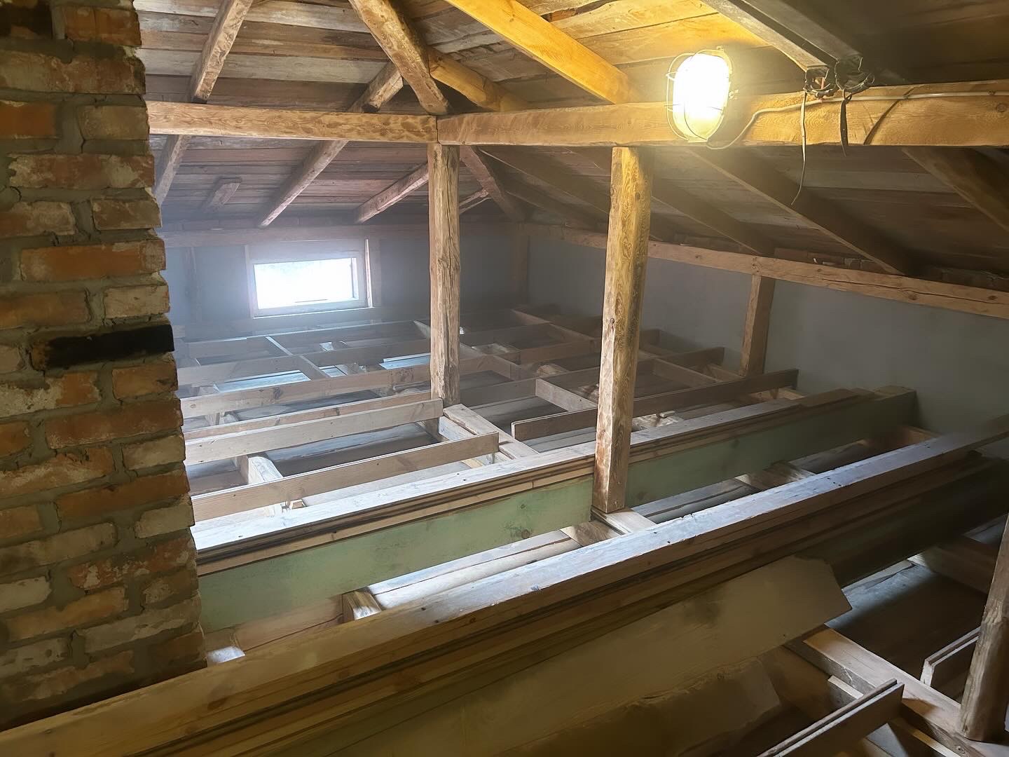 Poddasze w trakcie adaptacji: widoczne drewniane legary, ceglany komin oraz okno w szczycie, oświetlone lampą roboczą.