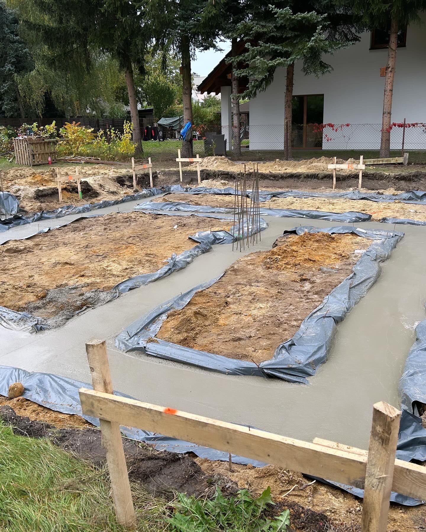 Wylany beton na przygotowane ławy fundamentowe z widocznym zbrojeniem i folią ochronną, teren budowy z drewnianymi znacznikami.