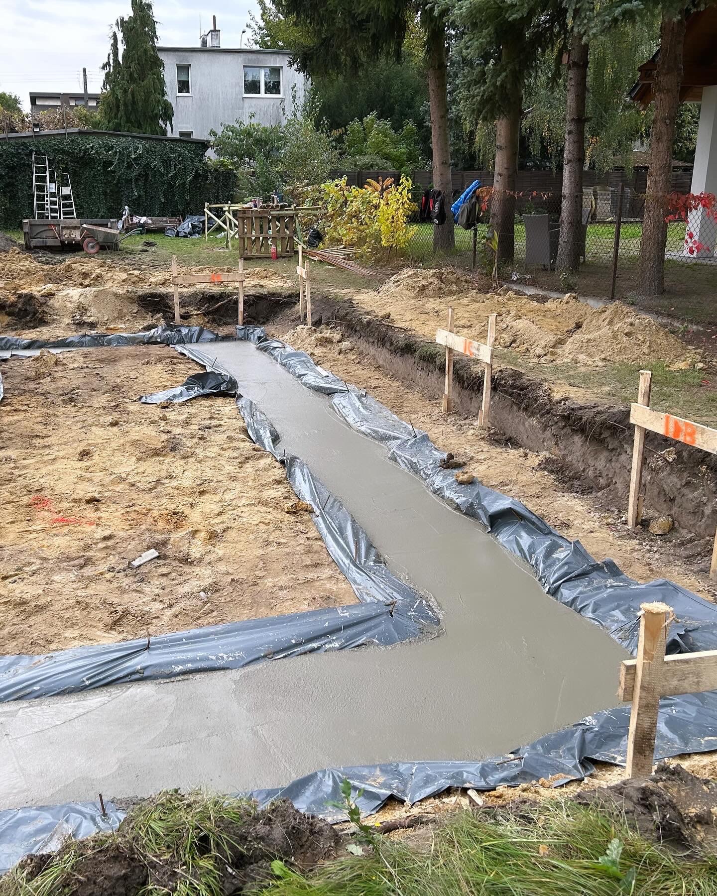 Wylany betonowy fundament pod budowę domu, zabezpieczony folią, widoczne drewniane szalunki i wykop, w tle drzewa i budynek mieszkalny.