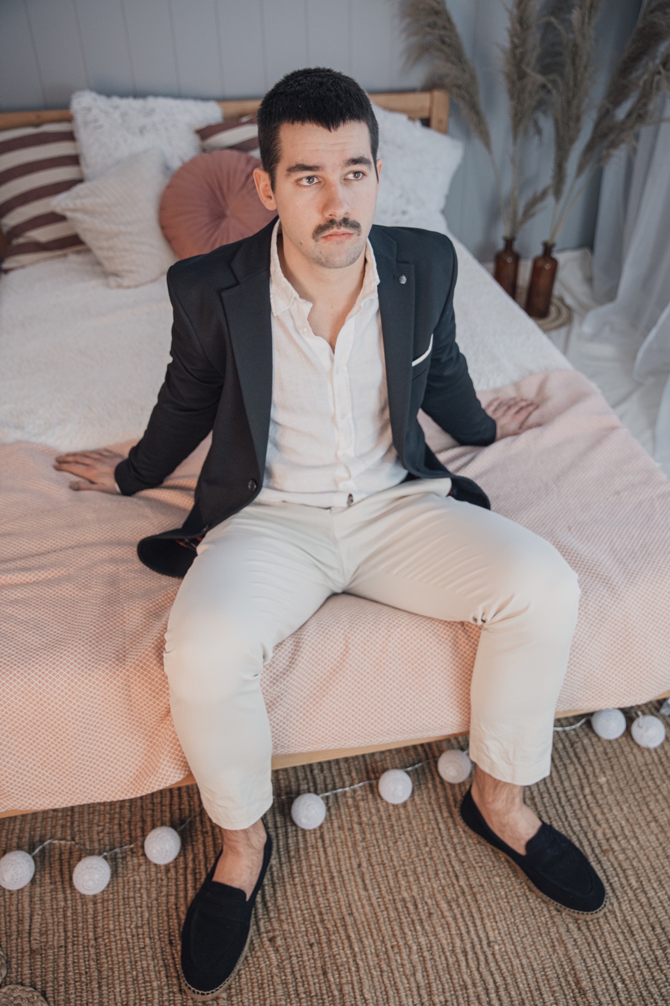 Mężczyzna z wąsami w rozpiętej białej koszuli i marynarce siedzi na łóżku z dekoracyjnymi poduszkami i lampkami cotton balls.