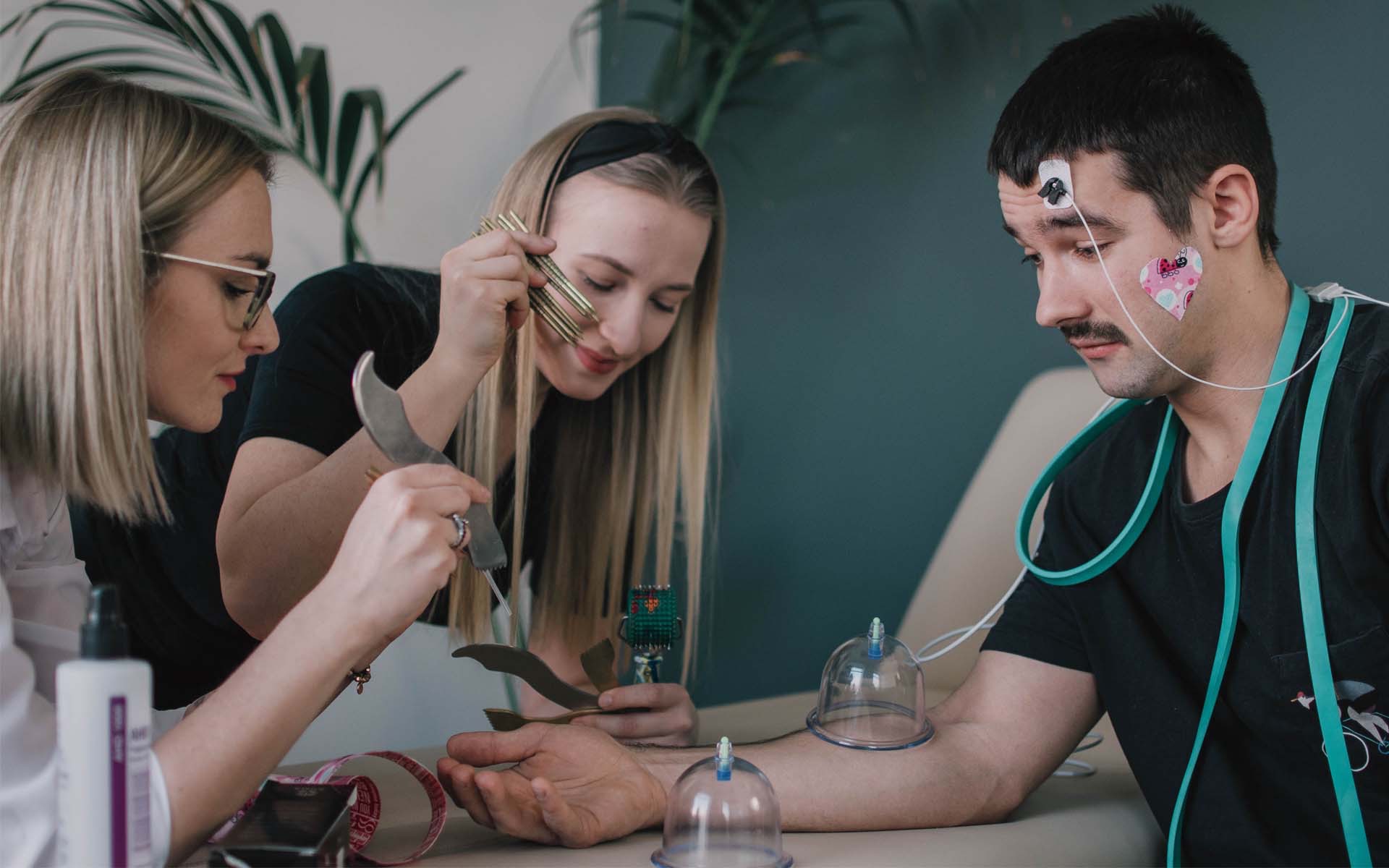 Mężczyzna z elektrodami i bańkami na ciele poddawany zabiegowi przez dwie kobiety używające metalowych narzędzi.