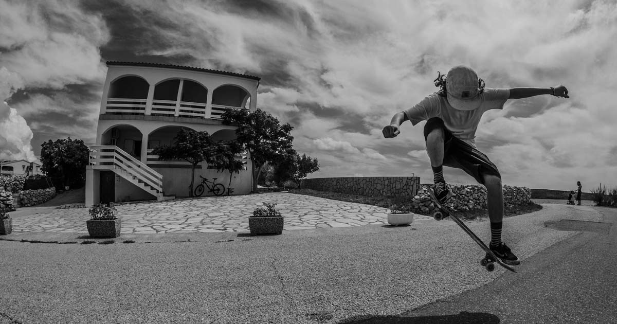 Czarno-białe zdjęcie młodego mężczyzny w locie na deskorolce przed białym budynkiem z balkonami i kamienną ścieżką, dynamiczne ujęcie z nisko wiszącymi chmurami.