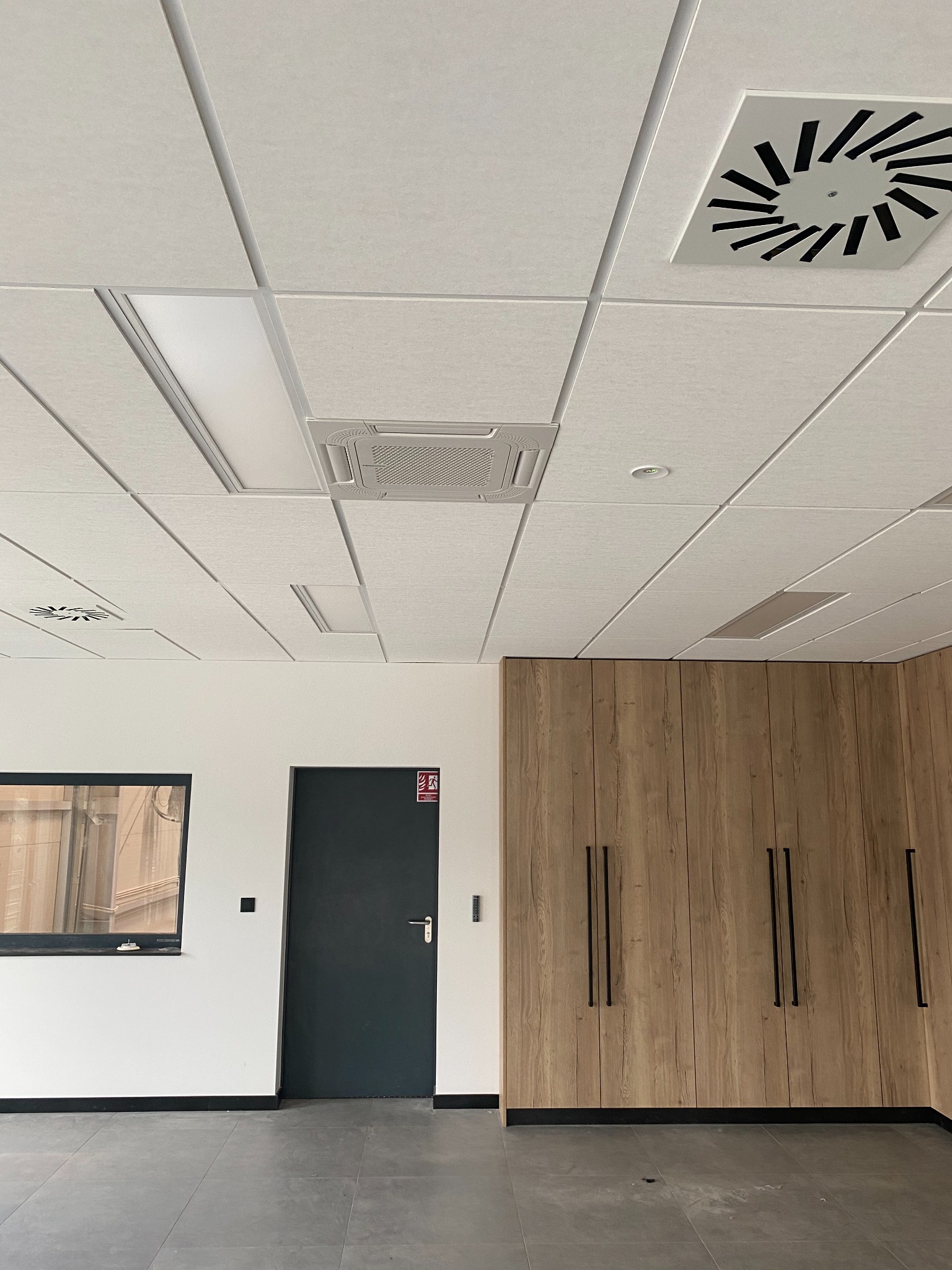 Sufit podwieszany w biurze z kratką wentylacyjną, panelem klimatyzacyjnym kasetonowym i oświetleniem LED, widok na ścianę z drzwiami i szafą w zabudowie.