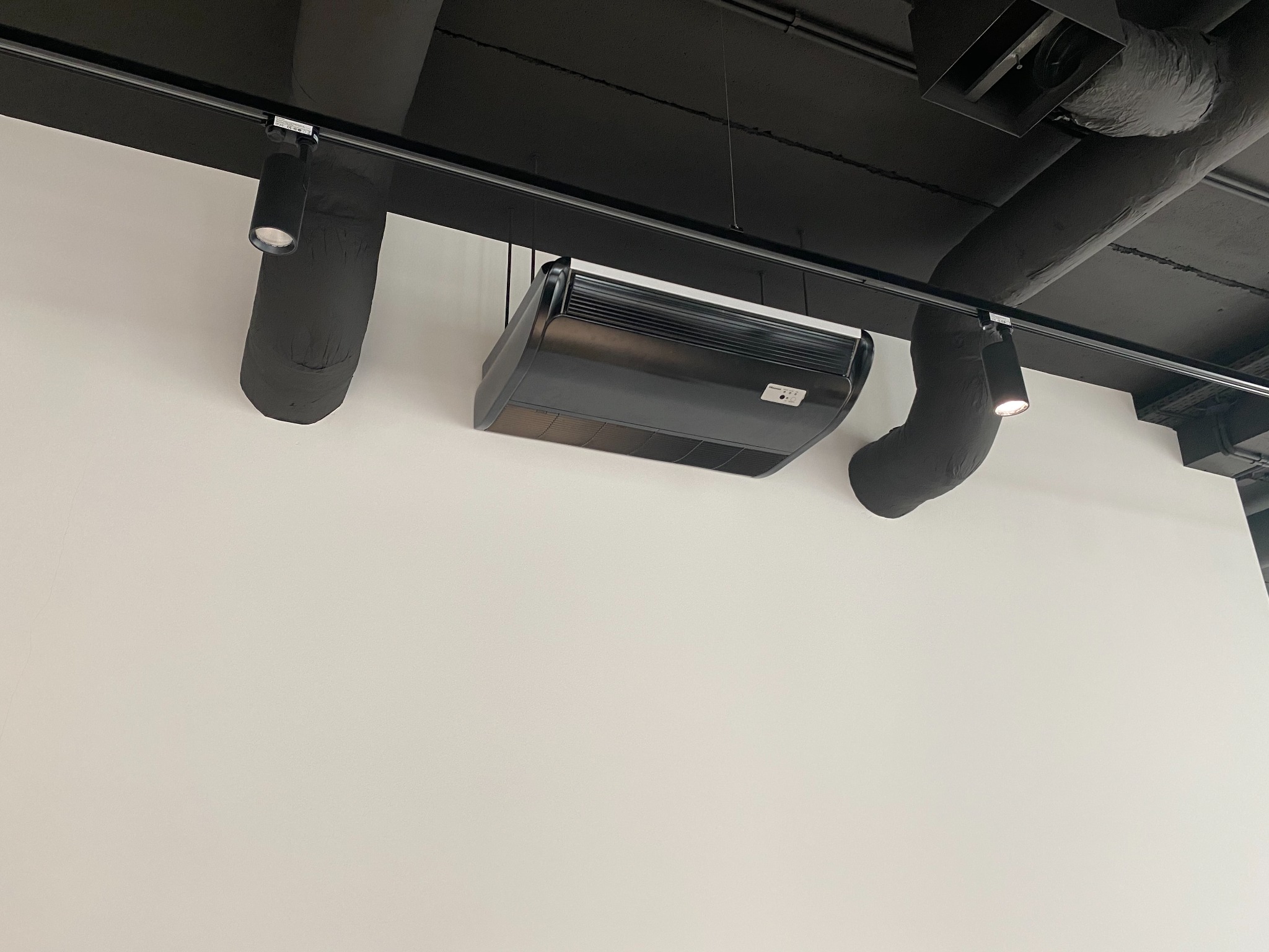 Czarna klimatyzacja kasetonowa zamontowana pod czarnym sufitem z widocznymi rurami wentylacyjnymi i oświetleniem punktowym.