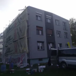 Wielopiętrowy budynek w trakcie ocieplania elewacji, widoczne rusztowania z siatką ochronną z nadrukiem reklamowym, częściowo pomalowana elewacja w odcieniach szarości, puszki z farbą i samochód...