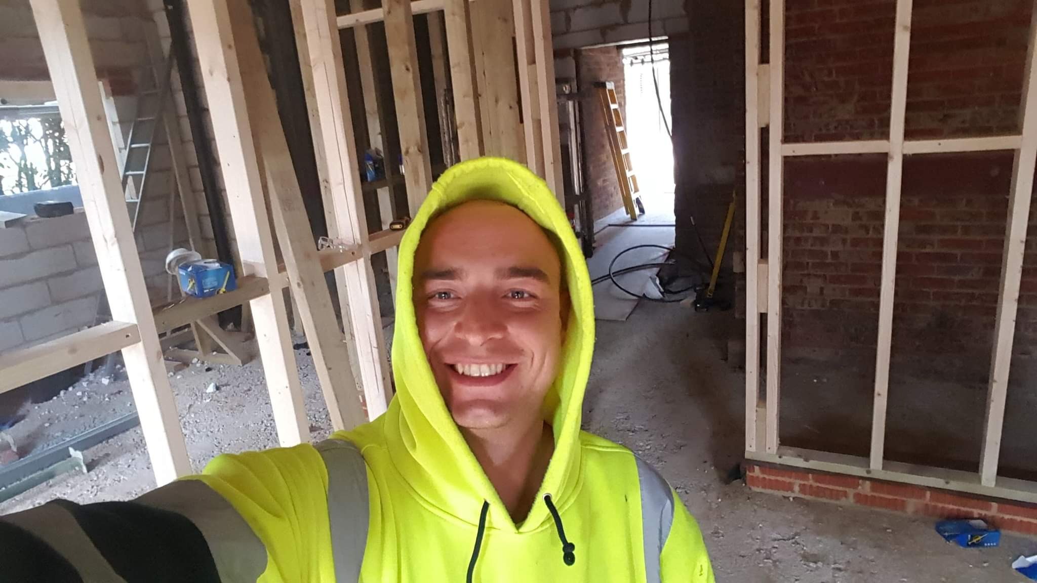 Uśmiechnięty pracownik w neonowej bluzie z kapturem robi selfie na tle konstrukcji ścian działowych z drewna i cegły w surowym stanie.