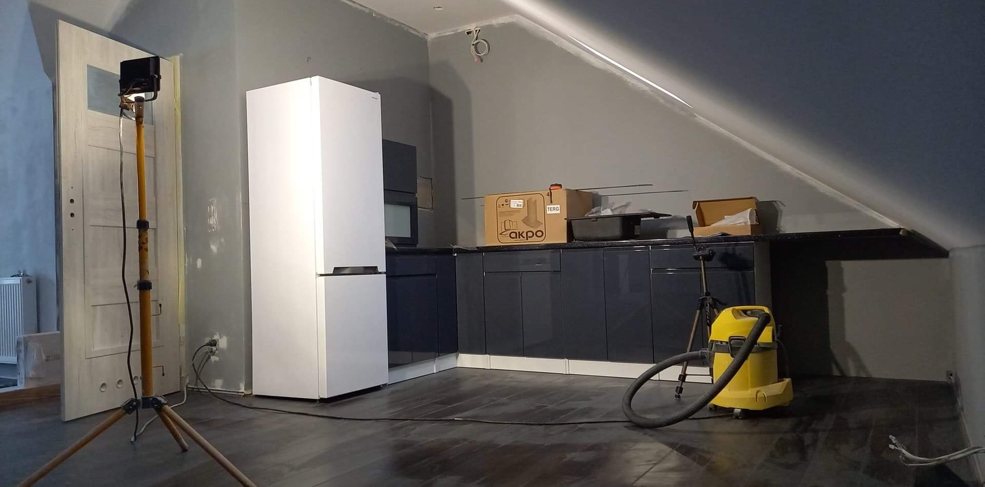Poddasze w trakcie wykańczania: widoczna zabudowa kuchenna z szafek w ciemnym kolorze, biała lodówka, lampa budowlana na statywie, odkurzacz przemysłowy oraz surowe ściany przygotowane do dalszych...