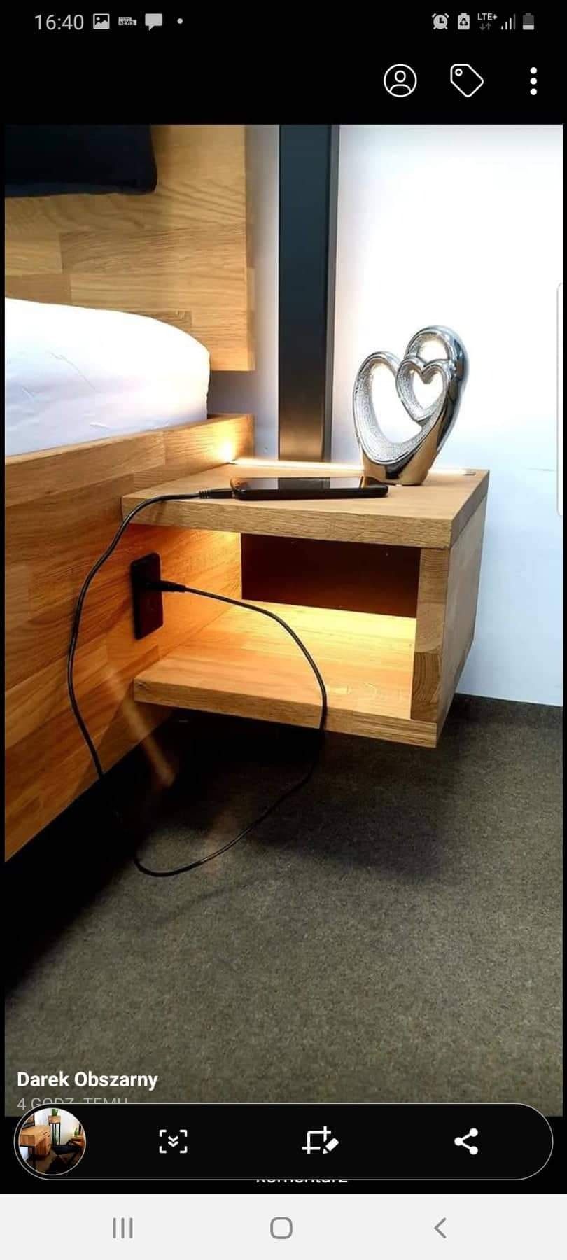 Drewniana szafka nocna, zamontowana przy drewnianym łóżku, z wbudowanym gniazdkiem USB i ładowarką do telefonu, oświetlona od spodu, z dekoracyjną figurką w kształcie serca na blacie.