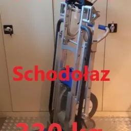 Schodołaz transportowy, srebrna konstrukcja z niebieskimi elementami na kółkach, widoczne naklejki z informacjami o wynajmie i wadze maksymalnej, stoi na metalowej podłodze.