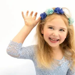 Portret uśmiechniętej dziewczynki z blond włosami i wiankiem z błękitnych kwiatów, ubrana w błękitną koronkową sukienkę, na białym tle.