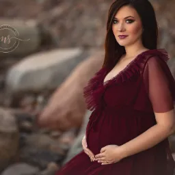 Kobieta w bordowej sukni z falbanami, trzymająca dłonie na brzuchu, pozuje na tle kamieni