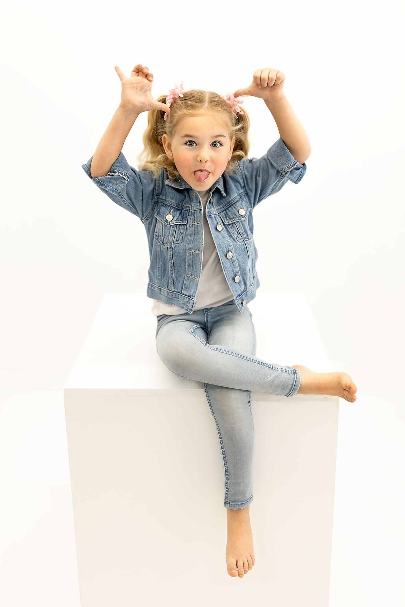Dziewczynka z blond włosami spiętymi w kucyki z różowymi kwiatkami, ubrana w jeansową kurtkę i spodnie, siedzi na białym sześcianie, wystawia język i robi rogi z palców na białym tle.