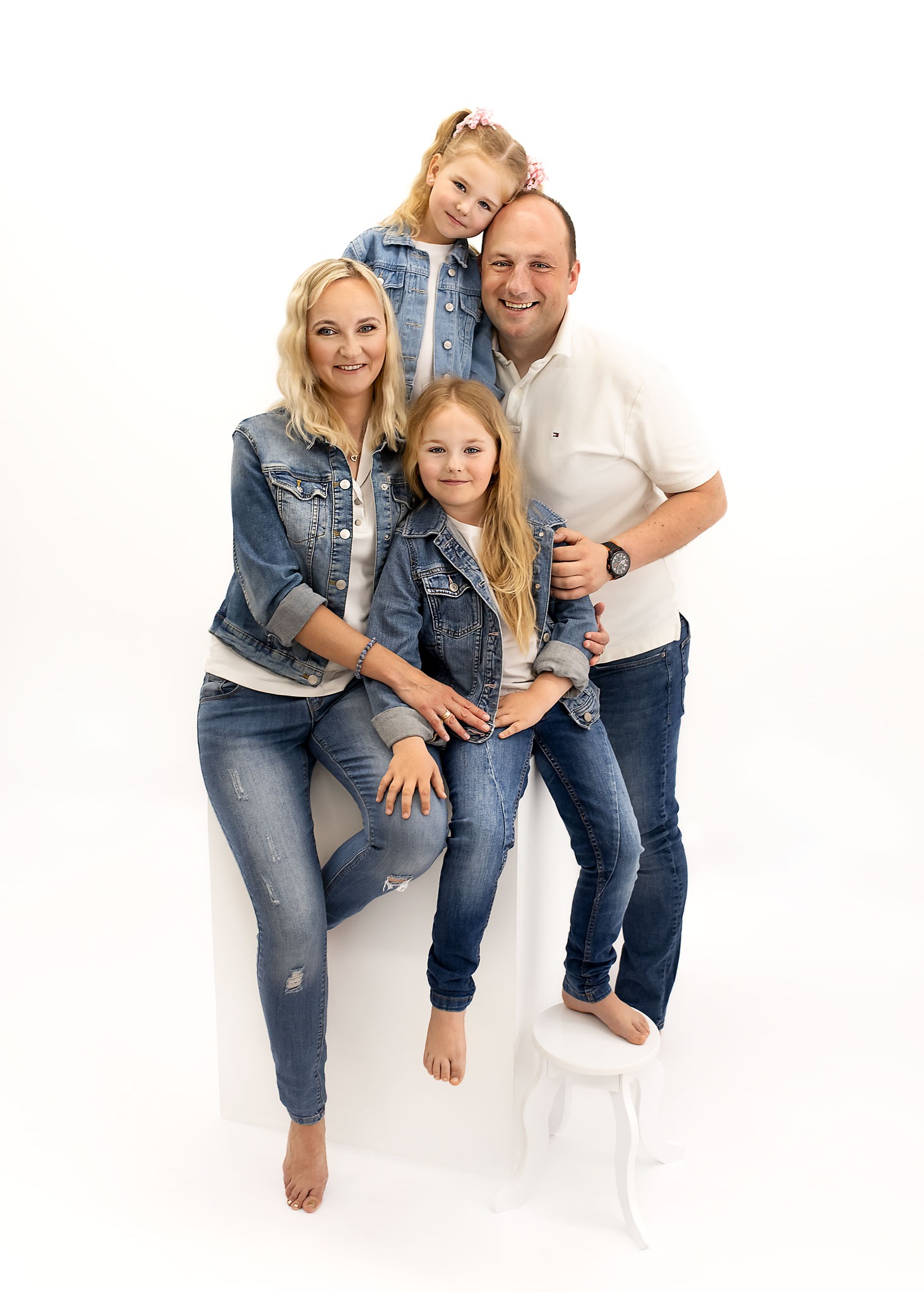Rodzinne portretowe ujęcie: mama siedzi boso na białym sześcianie, tata stoi za nią, obejmując dwie córki w jeansowych kurtkach na białym tle.