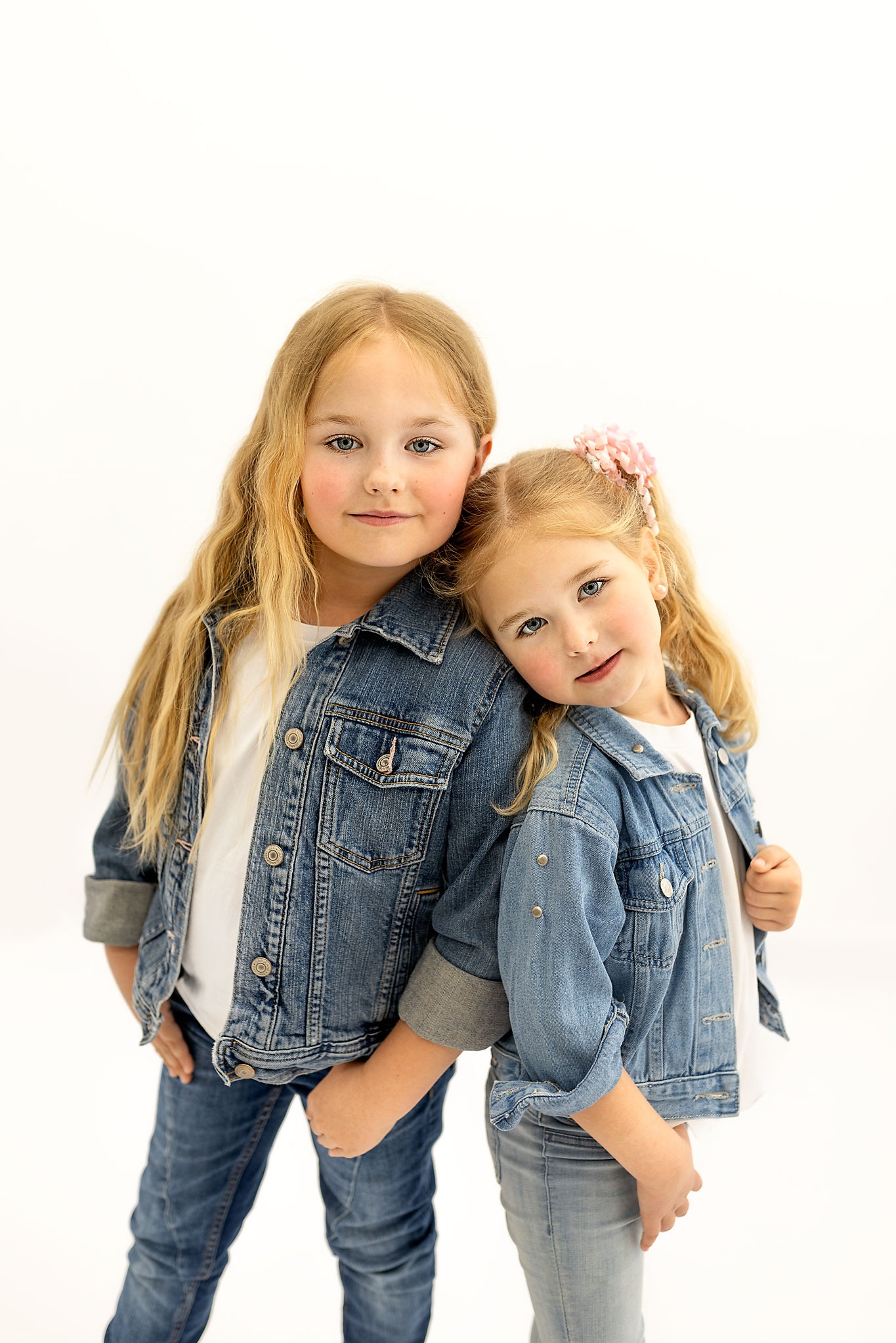 Dwie urocze dziewczynki, blondynki, w jeansowych kurtkach i spodniach, pozują na białym tle, jedna opiera głowę o ramię drugiej, patrząc w obiektyw.
