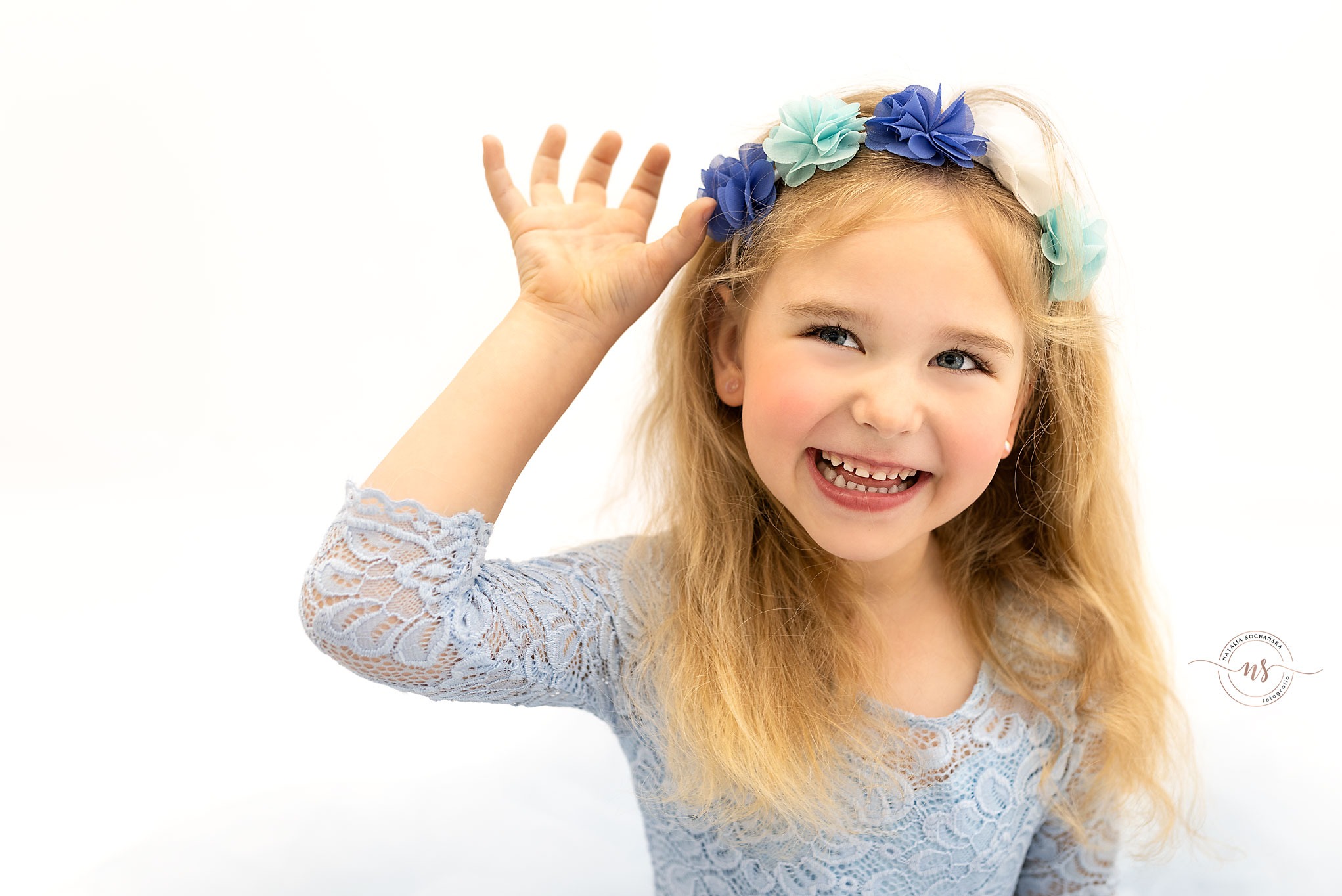 Portret uśmiechniętej dziewczynki z blond włosami i wiankiem z błękitnych kwiatów, ubrana w błękitną koronkową sukienkę, na białym tle.